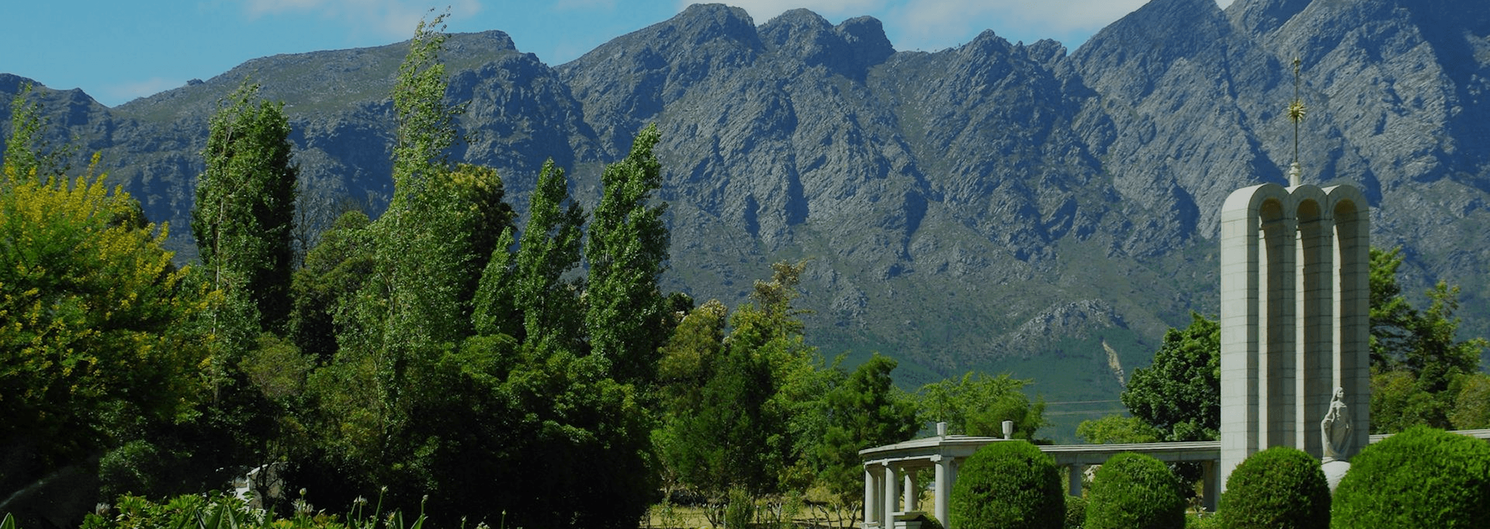 Jardín con árboles, montañas y un monumento bajo un cielo azul.
