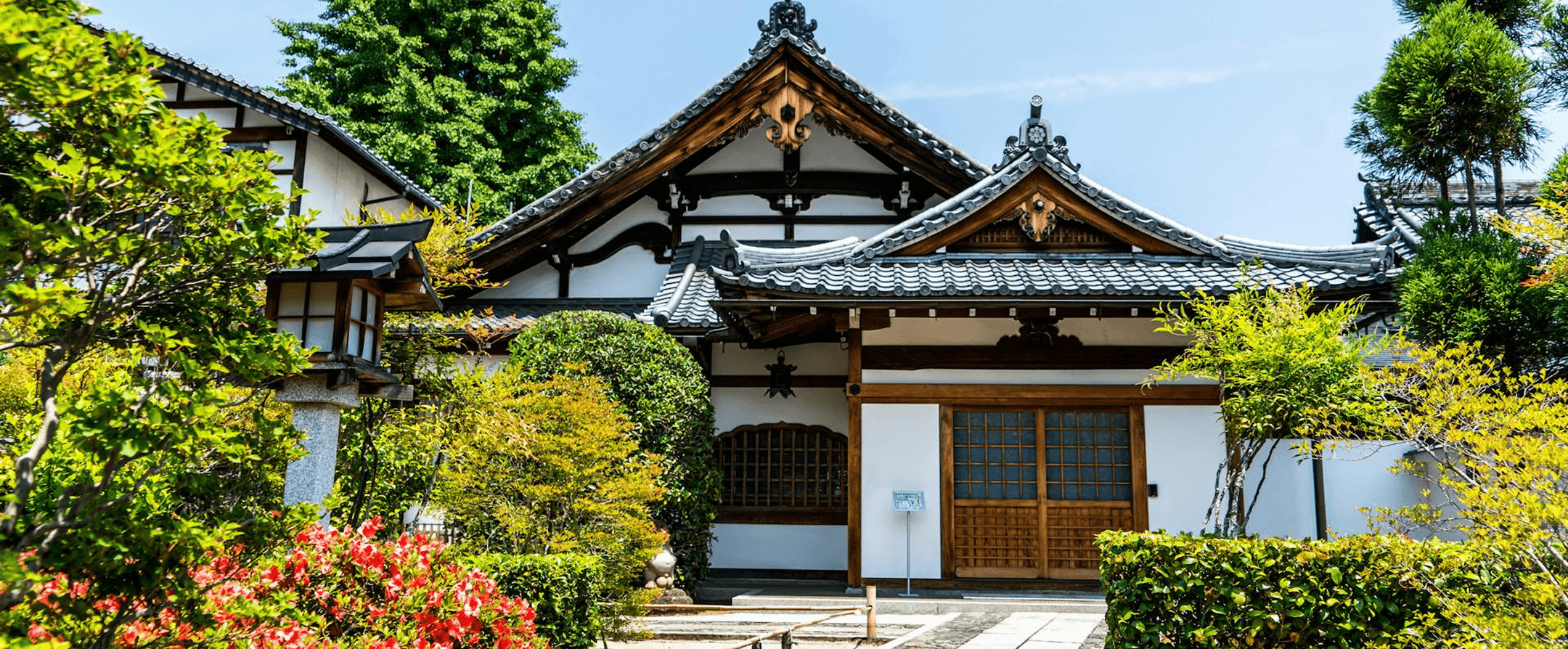 Templo japonés tradicional con techos de tejas curvadas y puertas correderas de madera, rodeado de exuberantes jardines verdes y arces en primavera